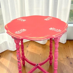 May include: A round, scalloped-edge side table painted in a vibrant coral-pink hue. The tabletop features four white floral stencils, while the four turned legs are finished in a bright fuchsia with visible distressed edges and a cross-brace base.