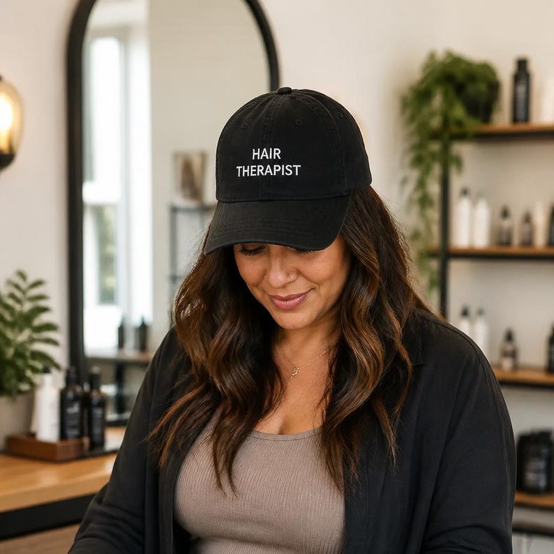 May include: A black baseball cap featuring the white embroidered text HAIR THERAPIST centered on the front panel. The cap has a structured crown, a curved brim, and a fabric strap closure at the back, providing a classic and simple design.