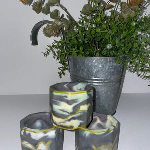 May include: Three small, hexagonal concrete planters featuring a marbled pattern of dark gray, white, and light green, with bright yellow painted edges along the top rims. The planters are arranged in a cluster, showcasing their geometric shape.