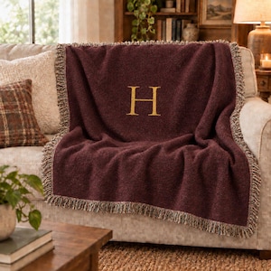 Op de afbeelding: Een diepbordeauxrode geweven plaid met een grote, centraal geplaatste gouden letter H. De rechthoekige deken heeft een getextureerd oppervlak en is afgewerkt met decoratieve franjes langs alle vier de randen voor een klassieke en gestructureerde uitstraling.