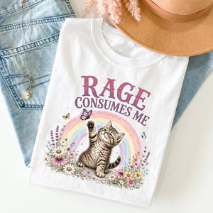 Puede incluir: Una camiseta blanca con el gráfico de un gatito atigrado alcanzando una mariposa morada. El diseño incluye un arcoíris pastel, ramas de lavanda y flores silvestres coloridas. El texto RAGE CONSUMES ME aparece en una fuente morada arqueada.