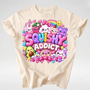 Op de afbeelding: Een cr&egrave;mekleurig t-shirt met een kleurrijke grafische print met de tekst SQUISHY ADDICT. Het ontwerp bevat stripfiguren zoals een aardbei, avocado, cupcake en melkpak, omringd door hartjes, sterren en een roze strik.