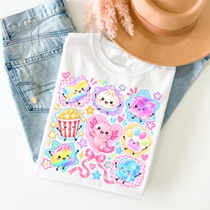 Op de afbeelding: Een wit t-shirt met een kleurrijke grafische print van een roze axolotl, een emmer popcorn, diverse pastelkleurige dumplings en geometrische vormen met lachende gezichten. Het ontwerp bevat kleine hartjes, sterretjes en een roze lintmotief.