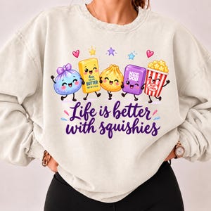 Op de afbeelding: Een beige sweater met ronde hals en een afbeelding van vijf stripfiguren, waaronder een blauw zakje, een gele boterstaaf met het label 4oz. Salted Butter, een knoedel, een paars blok met het label Nee Doh en popcorn. De tekst luidt: Life is better with squishies.