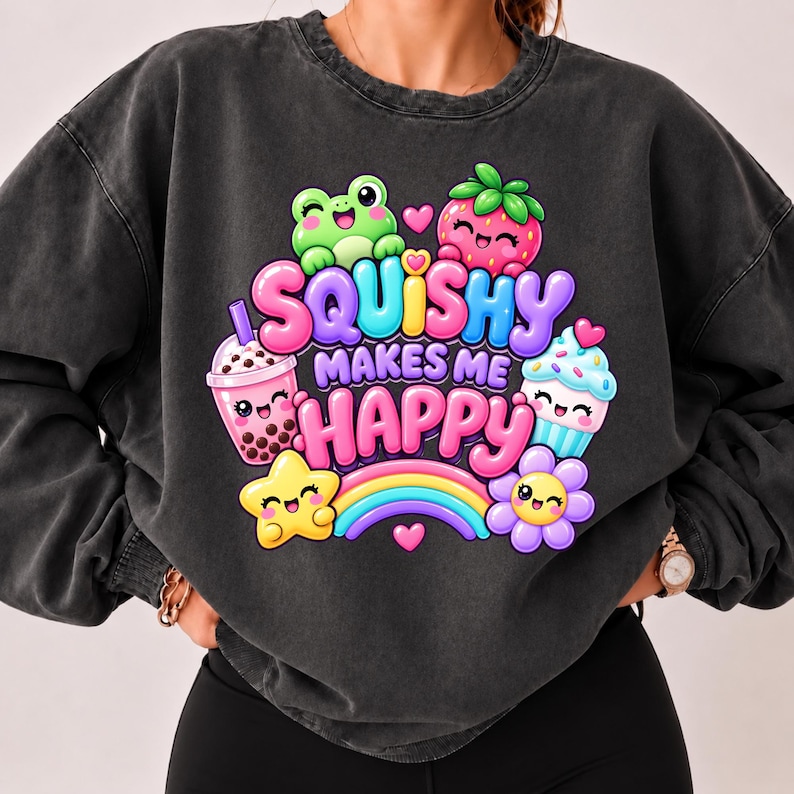 Op de afbeelding: Een antracietgrijze sweater met ronde hals en een kleurrijke grafische print met de tekst SQUISHY MAKES ME HAPPY. Het ontwerp bevat een groene kikker, een aardbei, een boba-thee, een cupcake, een gele ster, een regenboog en een paarse bloem.