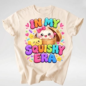 Op de afbeelding: Een cr&egrave;mekleurig t-shirt met een kleurrijke afbeelding van een lachende dumpling in een bamboe stoommandje met een roze strik, een blokje boter en de tekst "IN MY SQUISHY ERA" omringd door kleine hartjes, sterretjes en spikkels.