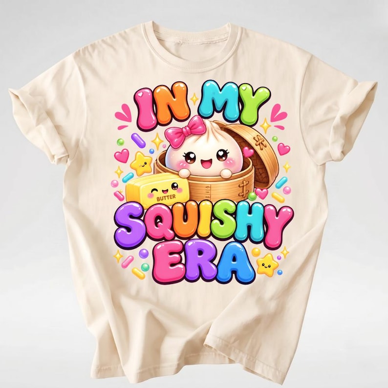 Op de afbeelding: Een cr&egrave;mekleurig t-shirt met een kleurrijke afbeelding van een lachende dumpling in een bamboe stoommandje met een roze strik, een blokje boter en de tekst "IN MY SQUISHY ERA" omringd door kleine hartjes, sterretjes en spikkels.
