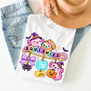 Op de afbeelding: Een wit t-shirt met een afbeelding van twee roze axolotls met heksenhoeden, waarvan er &eacute;&eacute;n een pompoen vasthoudt. Het ontwerp bevat een grote veiligheidsspeld met de tekst SQUISHIES, een paarse vichy-strik, een spin, een dumpling en een blok met het label NEE DOH SALTED BUTTER.