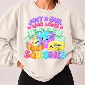 Op de afbeelding: Een beige sweater met ronde hals en een grafische print met de tekst "JUST A GIRL WHO LOVES SQUISHIES". Het ontwerp bevat een regenboog-dumpling in een bamboe stoommandje, een blauwe kubus en een blokje gezouten boter met "113 g".