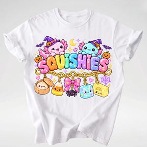 Op de afbeelding: Een wit t-shirt met een kleurrijke afbeelding van twee axolotls met heksenhoeden boven het woord SQUISHIES in bubbelletters. Daaronder hangt een gouden ketting met bedeltjes zoals een dumpling, maissnoepje, roze strik, spin, kubus en een blokje boter.
