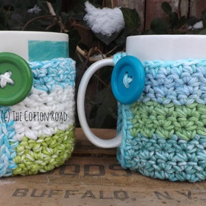 May include: Two white ceramic mugs with crocheted covers. The covers are in shades of blue, green, and white. The mugs have buttons attached to the covers.