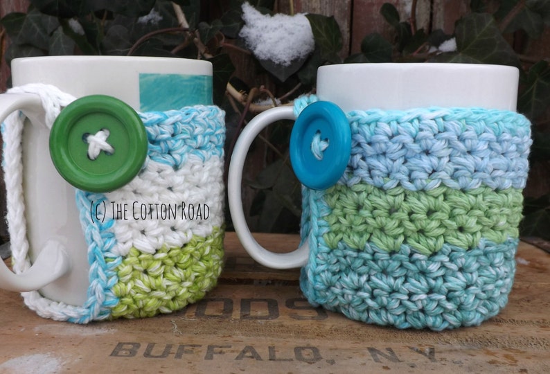 May include: Two white ceramic mugs with crocheted covers. The covers are in shades of blue, green, and white. The mugs have buttons attached to the covers.