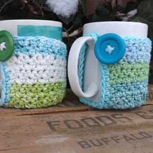 May include: Two white ceramic mugs with crocheted cup cozies. One cozy is white, green, and blue. The other is white, light blue, and green. Both cozies have a button on the front.
