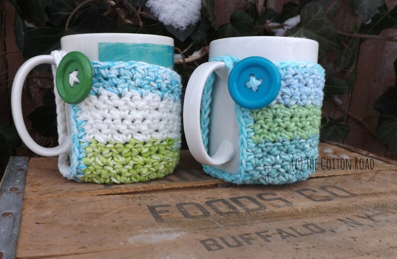 May include: Two white ceramic mugs with crocheted cup cozies. One cozy is white, green, and blue. The other is white, light blue, and green. Both cozies have a button on the front.