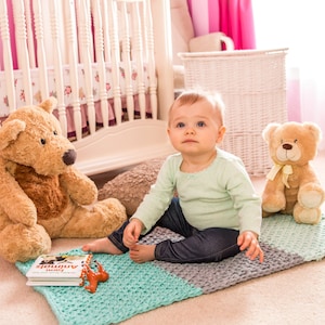 May include: A baby sits on a blue and gray knitted blanket with two teddy bears. The baby is wearing a light green long-sleeved shirt and blue jeans. The baby is looking at the camera. There is a book titled "First Animals" on the blanket.