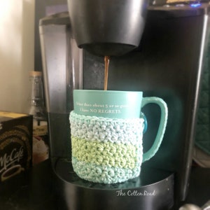 May include: A teal mug with the text "Other than about 5 or so guys I have NO REGRETS." is being filled with coffee from a coffee maker. The mug is wrapped in a crocheted green and blue mug cozy.