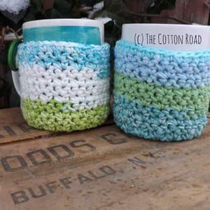 May include: Two crocheted mug cozies in blue, green, and white stripes. The cozies are on top of a wooden crate with the words "Air Foods Buffalo, NY" printed on it. (c) The Cotton Road