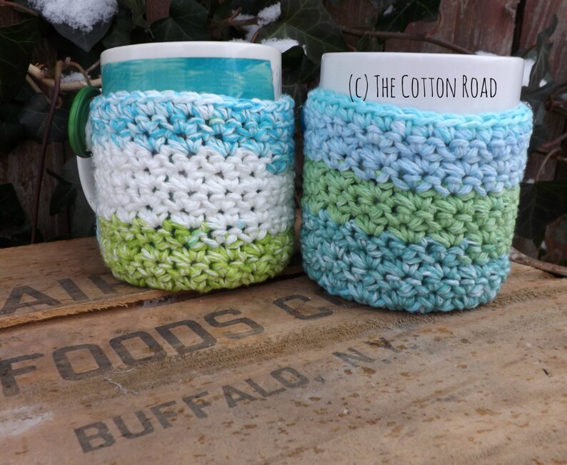 May include: Two crocheted mug cozies in blue, green, and white stripes. The cozies are on top of a wooden crate with the words "Air Foods Buffalo, NY" printed on it. (c) The Cotton Road