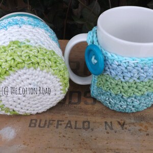 May include: Two crocheted mug cozies in green, blue, and white stripes. The cozies are made with a textured stitch and are designed to fit standard-sized mugs.