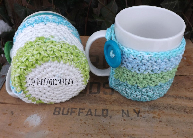May include: Two crocheted mug cozies in green, blue, and white stripes. The cozies are made with a textured stitch and are designed to fit standard-sized mugs.