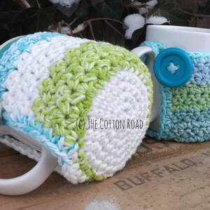 May include: Two crocheted mug cozies in a green, white, and blue color scheme. The cozies are made with a simple stitch pattern and feature a button on the handle.