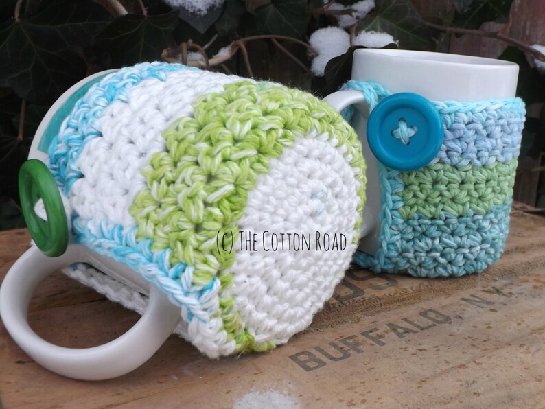 May include: Two crocheted mug cozies in a green, white, and blue color scheme. The cozies are made with a simple stitch pattern and feature a button on the handle.