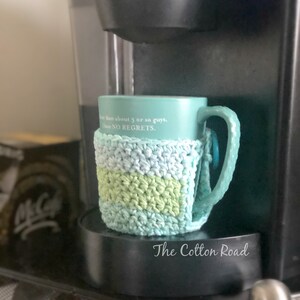 May include: A teal blue mug with the text "more than about 5 or so guys, I have NO REGRETS."  The mug is wrapped in a crocheted green and blue mug cozy. The cozy has a light blue, green, and white striped pattern. The mug is sitting on a black base with the text "The Cotton Road" printed on it.