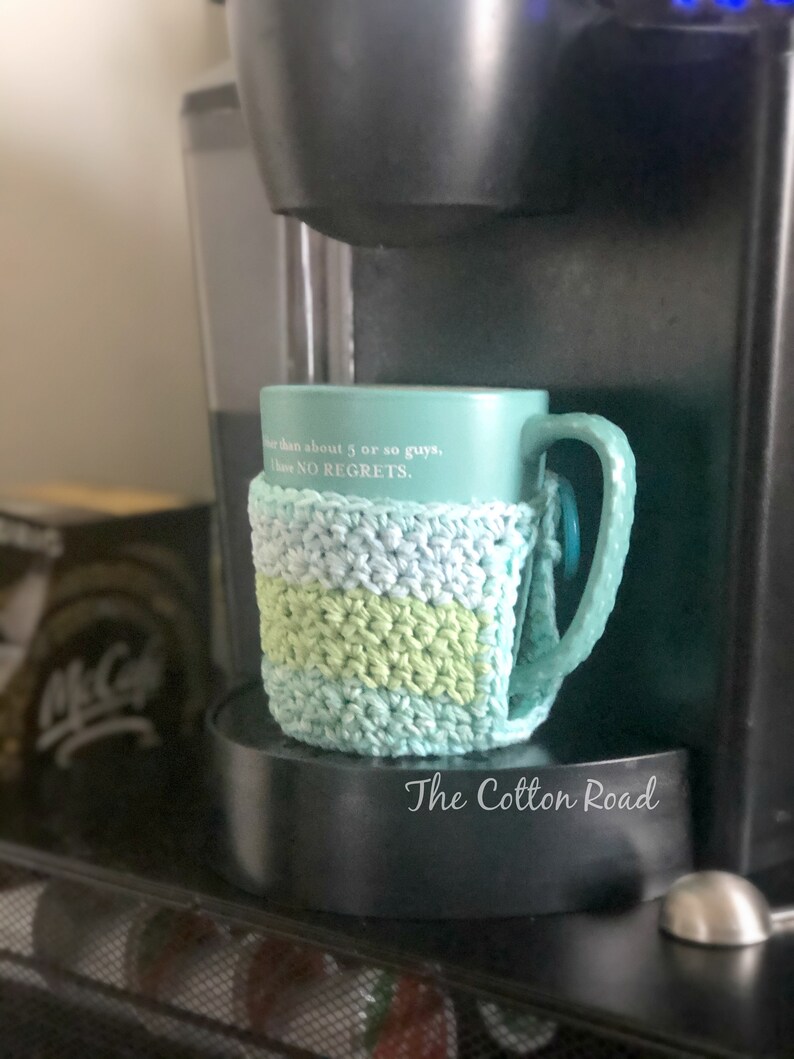 May include: A teal blue mug with the text "more than about 5 or so guys, I have NO REGRETS."  The mug is wrapped in a crocheted green and blue mug cozy. The cozy has a light blue, green, and white striped pattern. The mug is sitting on a black base with the text "The Cotton Road" printed on it.