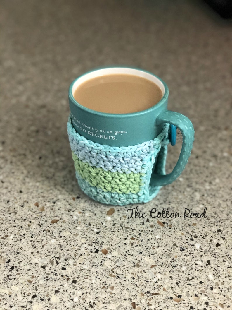 May include: A teal mug with a crocheted cozy in blue and green. The mug has the text "Been about 5 or so guys, NO REGRETS." on the side. The cozy has a green stripe. The mug is sitting on a speckled countertop. The text "The Cotton Road" is visible below the mug.
