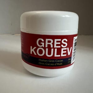 Puede incluir: Un tarro cilíndrico de plástico blanco con una etiqueta roja que presenta el texto blanco GRES KOULEV. Debajo, la etiqueta dice Haitian Gres Cacao y Gres Cacao d'Haiti, con un peso neto de 56g impreso en el lateral junto a un código de barras.