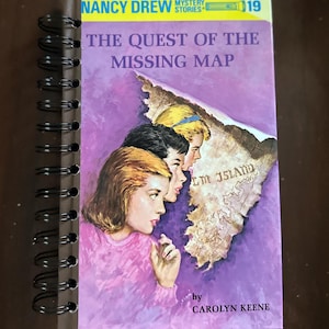 May include: A spiral-bound book titled Nancy Drew Mystery Stories 19, The Quest of the Missing Map by Carolyn Keene. The cover features a purple background with an illustration of three people looking at a map labeled LM Island.