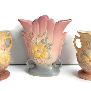 May include: Three vintage ceramic vases with floral designs. The vases are in shades of pink, yellow, and brown. The largest vase has a pink and white glaze with a yellow flower design. The two smaller vases have a yellow and brown glaze with a pink flower design.