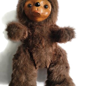 May include: A handmade brown plush toy with a wooden face. The toy has a furry body, arms, and legs. The face is carved with large black eyes, a small nose, and a smiling mouth. The toy is standing upright against a white background.