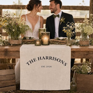 Op de afbeelding: Een beige linnen tafelloper met de zwarte opdruk THE HARRISONS EST. 2026. De loper ligt op een houten tafel met glazen kaarsenhouders en vazen gevuld met wilde bloemen, met twee personen zichtbaar op de achtergrond.