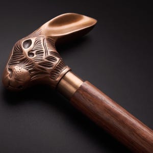 Può includere: Un bastone da passeggio con un'impugnatura in metallo color rame scolpita a forma di testa di coniglio con tratti del viso dettagliati e orecchie lunghe, fissata a un bastone in legno dritto color marrone scuro lucido con venature lisce.