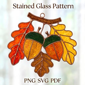 Op de afbeelding: Stained Glass Pattern PNG SVG PDF. Een decoratief hangend ontwerp met twee eikels met donkergroene kapjes en oranje lichamen, omringd door herfstkleurige eikenbladeren in rood, oranje en geel, omlijnd met koperkleurig metaal.