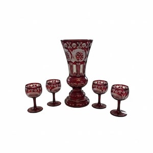 May include: A set of five pieces of ruby red cut-to-clear glass, comprising one large pedestal vase and four matching stemmed goblets. Each piece features intricate etched floral and geometric patterns that reveal clear glass beneath the red.