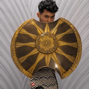 May include: A large, circular, metallic shield featuring a prominent central medallion with intricate floral engravings. The shield has a dark, weathered finish with radiating golden rays extending toward the scalloped, riveted outer edge.