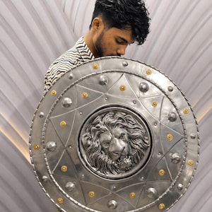 Puede incluir: Un escudo circular de metal grande que presenta un relieve tridimensional prominente de una cabeza de león en el centro. El escudo está decorado con una serie de tachuelas plateadas en relieve y pequeños acentos dorados en forma de estrella.