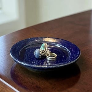 May include: A small, round, dark blue ceramic dish with a speckled texture. The dish holds three rings: one with a large blue stone, one with a turquoise stone, and a silver band. The dish sits on a dark wooden surface.