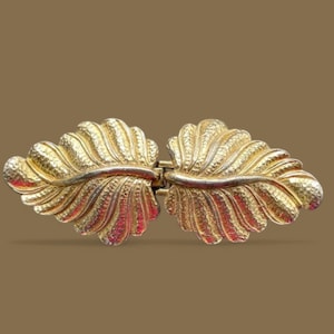 May include: A gold-colored clasp, featuring a leaf design. The clasp is composed of two symmetrical, textured leaf shapes connected by a hinge. The leaves have detailed veining and a hammered texture, creating a vintage aesthetic.