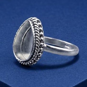 May include: A silver-toned ring featuring a teardrop-shaped bezel setting. The bezel is surrounded by a decorative beaded and twisted rope border. The band is smooth and plain, connecting to the base of the teardrop-shaped setting.