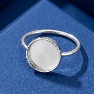 May include: A silver-toned metal ring featuring a circular, flat-bottomed bezel setting. The ring has a thin, smooth band and a polished, reflective surface on the circular tray, designed to hold a cabochon or other decorative inlay element.