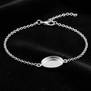 May include: A silver-toned metal bracelet featuring a delicate chain and a central oval-shaped bezel setting. The piece includes a small lobster claw clasp for closure, designed to hold a cabochon or inlay within the smooth, recessed metal frame.