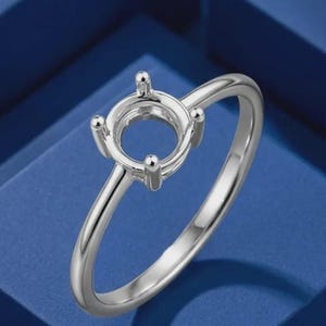 May include: A polished silver-toned metal ring setting featuring a four-prong head designed to hold a single gemstone. The band is smooth and slender, showcasing a classic solitaire style with a high-shine metallic finish and a circular mount.