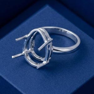 May include: A polished silver ring setting featuring an oval-shaped head with four prongs designed to hold a gemstone. The band is smooth and circular, showcasing a metallic finish that reflects light across its curved surface and prong structure.