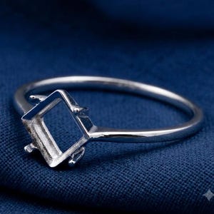May include: A silver-toned metal ring featuring a square-shaped setting designed to hold a gemstone. The ring has a thin, smooth band and four small prongs extending from the corners of the square frame to secure a stone in place.