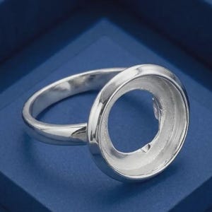 May include: A polished silver ring featuring a circular bezel setting with a smooth, reflective surface. The ring has a simple, rounded band that connects to the open, cup-like circular head, designed to hold a stone or decorative element.