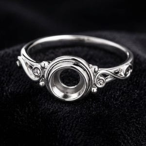 May include: A silver-toned metal ring setting featuring a circular bezel cup designed to hold a gemstone. The band displays intricate scrollwork and swirling filigree patterns on both sides of the central setting, creating a detailed metal structure.