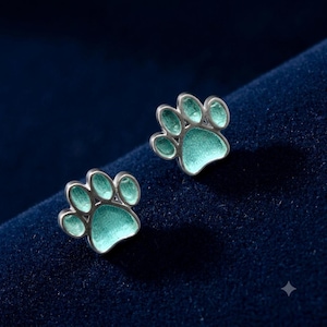 May include: A pair of silver-toned stud earrings shaped like animal paw prints. Each earring features a metallic silver border surrounding a textured, light mint green enamel inlay that fills the central pad and the four individual toe pads.